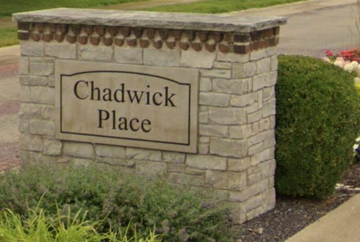 The stone entrance monument at Chadwick Place: a carved limestone panel reading
                  Chadwick Place, set into a low wall of stacked grey stone with a dentil course
                  beneath its cap, framed by clipped shrubs and flowers at the neighborhood entry.